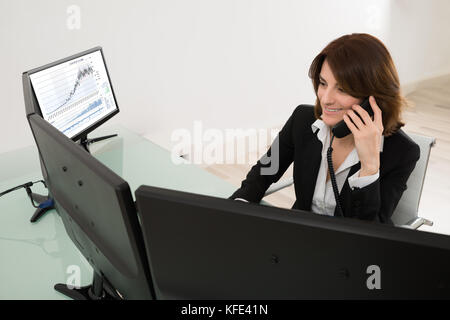 Sorridente giovane donna borsa broker lavora su più computer mentre si parla al telefono Foto Stock