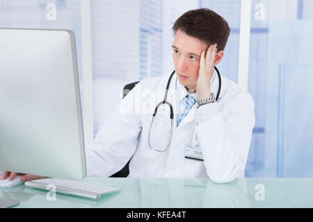 Preoccupato per il giovane maschio medico tramite il computer al banco in ospedale Foto Stock
