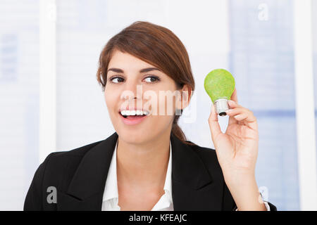 Ritratto di felice imprenditrice tenendo la luce verde lampadina in ufficio Foto Stock