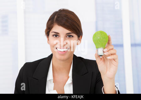 Ritratto di felice imprenditrice tenendo la luce verde lampadina in ufficio Foto Stock