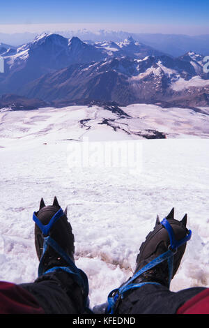 Prospettiva in prima persona di corpo e di gran lunga il paesaggio. L'uomo le gambe di ramponi prendere un periodo di riposo sulla cima della montagna. Piedi in scarpe da trekking sullo sfondo del Foto Stock
