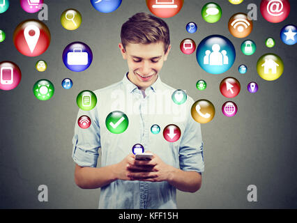 Tecnologia mobile high-tech. Giovane felice che utilizza uno smartphone con icone delle applicazioni di social media che volano fuori dallo schermo isolato su una parete grigia Foto Stock