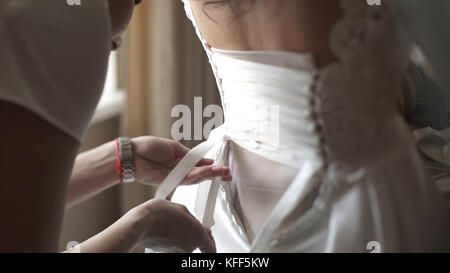 Eleganti e belle damigelle d'onore che aiutano la splendida sposa bruna in abito bianco a prepararsi per il matrimonio, i preparativi mattutini, la donna che indossa un abito. Mamma e damigella, aiutate a vestire un abito da sposa Foto Stock