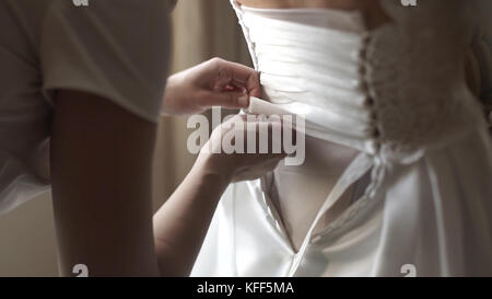 Eleganti e belle damigelle d'onore che aiutano la splendida sposa bruna in abito bianco a prepararsi per il matrimonio, i preparativi mattutini, la donna che indossa un abito. Mamma e damigella, aiutate a vestire un abito da sposa Foto Stock