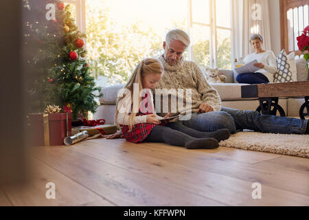 Bambina con il nonno seduto sul pavimento e con tavoletta digitale durante il Natale, con gran Madre seduti sul divano sul retro. Bambina di un Foto Stock