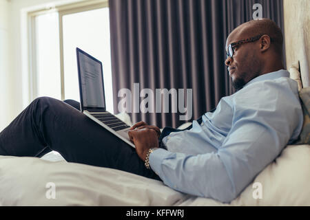 Uomo in abiti formali lavorando su laptop mentre giaceva a letto. Imprenditore lavora su computer portatile dalla sua camera d'hotel. Foto Stock