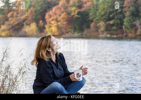 Felice donna seduta con caffè lungo il lago in autunno Foto Stock