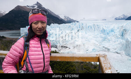 Giovani viaggiatori asiatici in piedi sul ghiacciaio passerella visitatori in camicia rosa e hat guardando la fotocamera con il Glacier e la gamma della montagna in background. Foto Stock