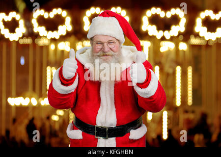 Il vecchio Babbo Natale dando pollice in alto con entrambe le mani. positivo babbo natale dando il pollice fino gesto su anno nuovo sfondo luci. realistico santa claus su Foto Stock