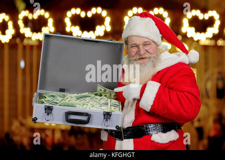 Santa Claus tenendo la valigia con il denaro. senior santa claus puntando con il dito indice sul centinaio di dollari nella valigetta, sfondo sfocato. dono su ch Foto Stock