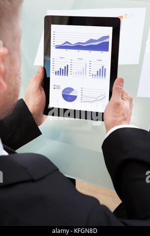 Imprenditore guardando una serie di grafici sul suo computer tablet come lavora al suo banco sopra la spalla vista dello schermo Foto Stock