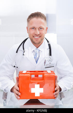 Sorridente medico o paramedico in un bianco camice con uno stetoscopio intorno al suo collo tenendo un riquadro rosso contenente un kit di primo soccorso Foto Stock