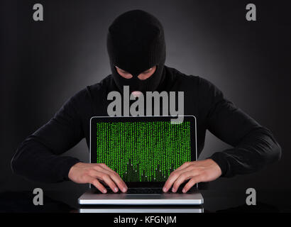 Hacker in un balaclava in piedi nel buio furtivamente rubare dati da un computer portatile o inserire spyware in una sicurezza online e rischio conce Foto Stock