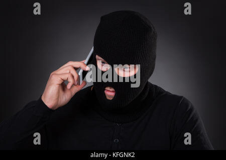 Ladro di un passamontagna e abito nero in piedi al buio utilizzando un furto di telefono cellulare o un terrorista attivando una bomba in remoto Foto Stock