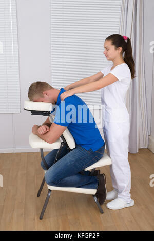 Fisioterapista femmina dando massaggio della spalla per uomo sulla poltrona di massaggio Foto Stock