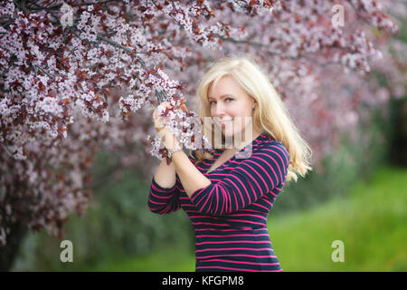 Sorridenti Caucasian donna bionda con capelli lunghi in viola fedora hat nei pressi di prugna fiore ciliegio, gode il fiore Foto Stock