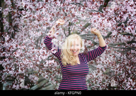 Sorridenti Caucasian donna bionda con capelli lunghi in viola fedora hat nei pressi di prugna fiore ciliegio Foto Stock