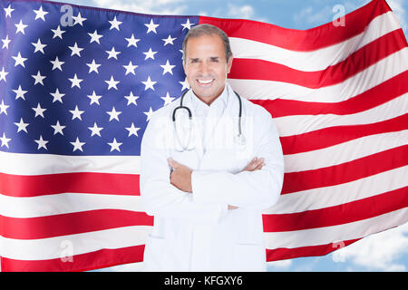 Ritratto di fiducioso maschio maturo medico in piedi nella parte anteriore della bandiera americana Foto Stock