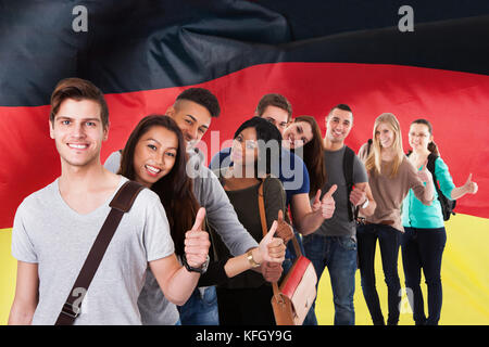Gruppo di studenti multietnici in piedi di fronte alla bandiera tedesca mostrando Thumb Up Foto Stock