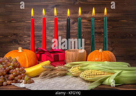 Festival kwanzaa concetto con sette candele rosso, nero e verde, confezione regalo, zucche, spighe di grano, uva, calli, banana, ciotola e frutti su legno Foto Stock