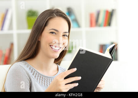 Felice ragazza del lettore di leggere un libro e guardando la telecamera seduto su un divano a casa Foto Stock