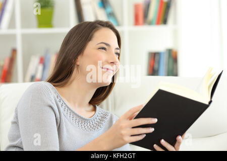 Donna sogna la lettura di un libro e guardando lontano seduto su un divano a casa Foto Stock