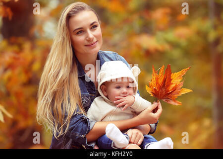 Ritratto di una giovane madre bionda con carino piccolo figlio seduto in autunno park su albero secco fogliame sfondo, famiglia felice con piacere la spesa Foto Stock