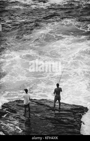 Due uomini stanno su una roccia in acque scoscese a pescare lungo la passeggiata costiera di Bondi fino a Coogee a Sydney, Australia. Foto Stock