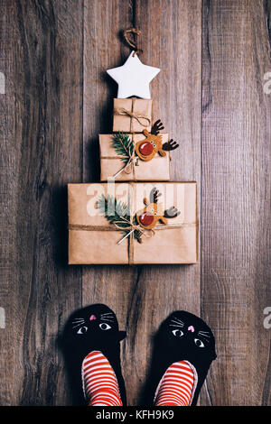 Tre marrone carta avvolta doni stabiliti nella forma di un albero di natale con stella sulla parte superiore. donna in piedi in cat pantofole, striped calze. vista dall'alto. Foto Stock