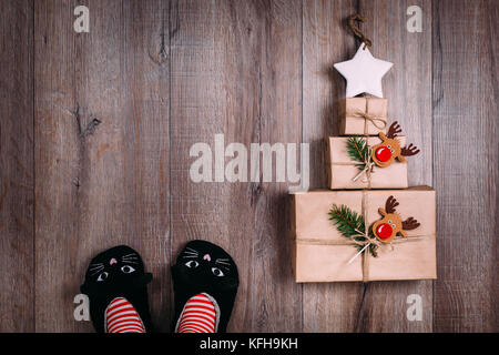 Tre marrone carta avvolta doni stabiliti nella forma di un albero di natale con stella sulla parte superiore. donna in piedi in cat pantofole, striped calze. vista dall'alto. copyspace Foto Stock