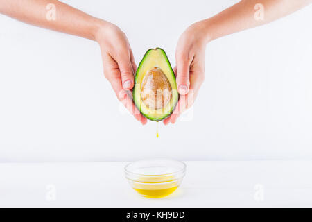 Fresh avocado con olio cadere in mani femminili e una ciotola di olio sul bianco sullo sfondo di legno isolata. sani e naturali di bellezza concetto. vista frontale. spac Foto Stock