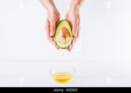 Fresh avocado con olio cadere in mani femminili e una ciotola di olio sul bianco sullo sfondo di legno isolata. sani e naturali di bellezza concetto. vista frontale. spac Foto Stock