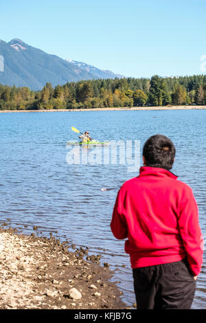 Un ragazzo asiatico in felpa rossa guarda un kayaker nelle calme acque del lago autunno giornata di sole Foto Stock