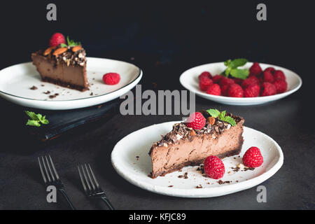 Due pezzi di cioccolato cheesecake decorate con lamponi e menta su piastra bianca su sfondo scuro. Foto Stock