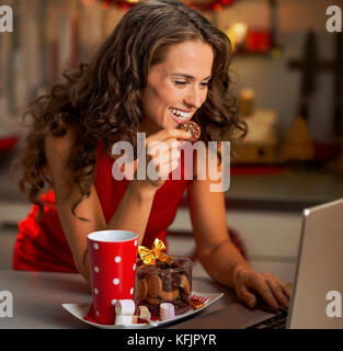 Felice giovane casalinga con spuntini di natale utilizzando laptop in cucina Foto Stock