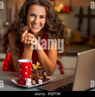 Felice giovane casalinga con spuntini di natale utilizzando laptop in cucina Foto Stock