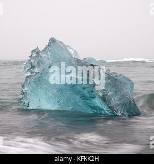 Iceberg blu turchese galleggiante in acque glaciali e onde in una giornata nuvolosa grigia vicino alla laguna glaciale di Jokulsarlon, Vatnajokull, Islanda del Sud Foto Stock