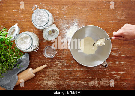 Mano con cucchiaio versare la farina in un recipiente di cottura sulla scena tavolo in legno Foto Stock