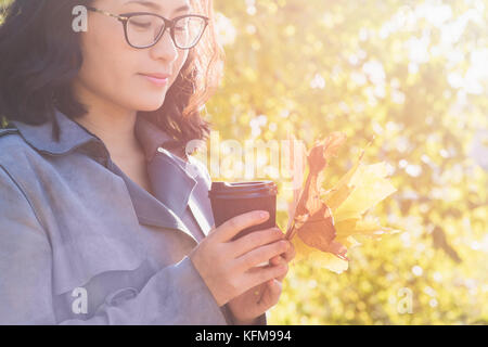 Bella giovane donna asiatica di bere una bevanda calda da carta monouso cup all'aperto nella bella fogliame di autunno. Foto Stock