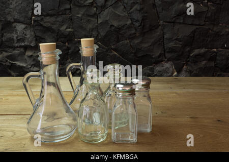 Olio d'oliva, aceto di bottiglie di vetro, il sale e il pepe impostato sul tavolo di legno, nero contro il muro di pietra Foto Stock