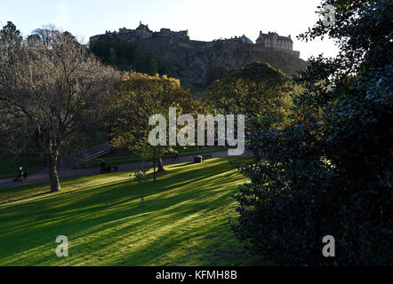 La luce solare bassa getta ombre attraverso gli alberi nei giardini di Princes street, con il Castello di Edimburgo stagliano in background. Foto Stock