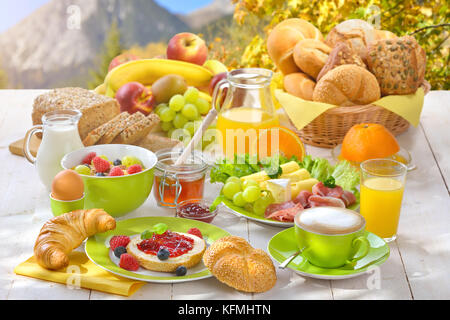 Fuori è servita la prima colazione con una tazza di cappuccino e una grande selezione di cibo, montagne e fogliame di autunno in background Foto Stock