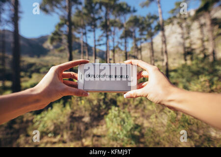 Mani tenendo una smart phone contro una foresta sfocata sullo sfondo. Telefono cellulare schermo che visualizza le parole di esplorare il mondo. Foto Stock