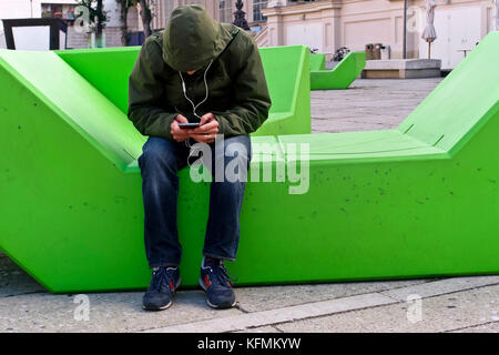 Giovane uomo caucasico irriconoscibile agganciato, volto nascosto, seduto su una moderna panchina a Museumsplatz, testando sul cellulare. Vienna, Austria, Europa UE Foto Stock