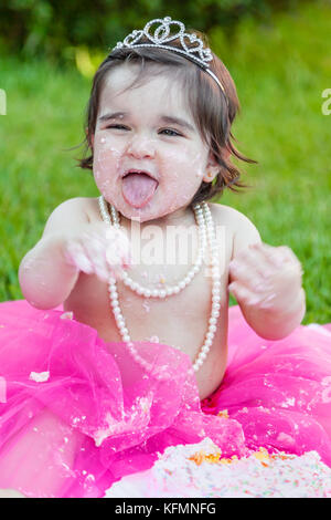 Sorridente happy baby toddler girl primo compleanno festa anniversario. ridendo e con la lingua fuori, faccia sporca da rosa torta. princess tiara costume Foto Stock