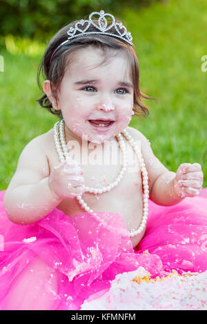 Sorridente happy baby toddler girl primo compleanno festa anniversario. a ridere con il viso e le mani sporche da fracassato torta rosa. princess tiara costume Foto Stock