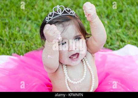 Felice eccitato toddler Baby girl primo compleanno festa anniversario. grida urrà con pugni sollevata in gioia. il viso e le mani sporche di fracassato torta rosa Foto Stock