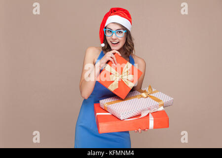 Giovane donna adulta in vestito blu e occhiali azienda molti confezione regalo, guardando la fotocamera e sorriso toothy. su sfondo marrone. studio shot Foto Stock