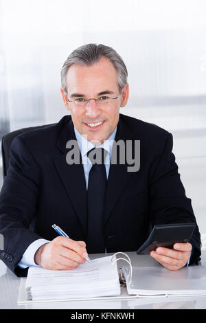 Happy businessman matura il calcolo di finanziare con la calcolatrice alla scrivania Foto Stock