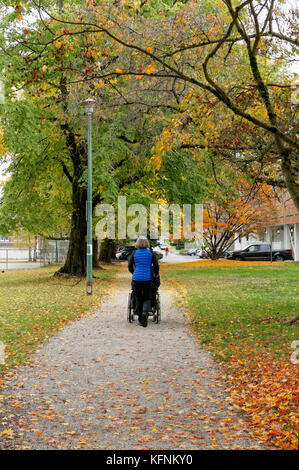 Mddle in età donna spingendo una persona anziana in una sedia a rotelle attraverso un parco in autunno, Vancouver, BC, Canada Foto Stock